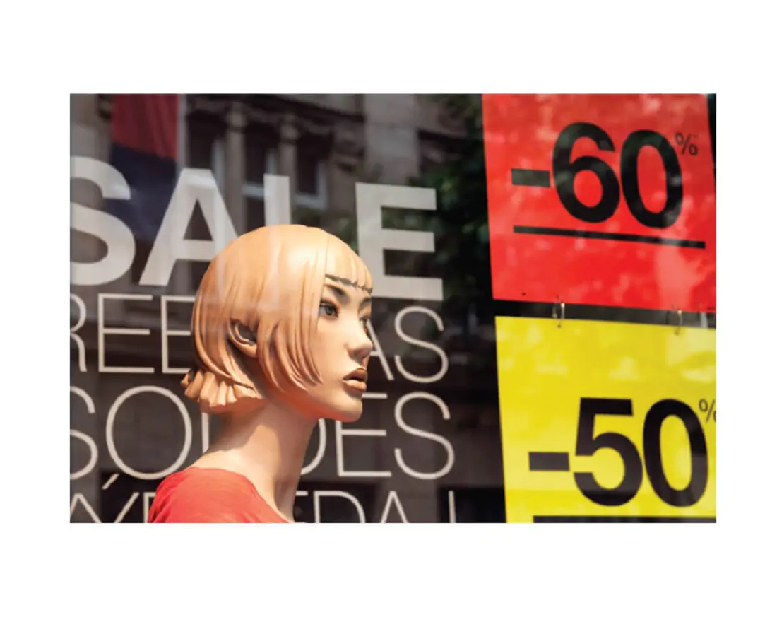 Women manikin standing in front of a store window with Sale on it and red and yellow signs with sale %