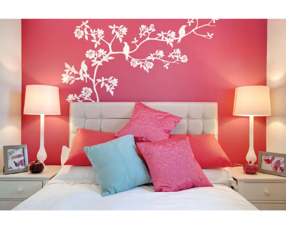 A bedroom with a pink wall and a flower branch, white bed with pink and blue pillows 