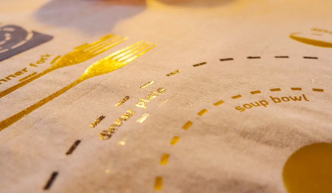 White cloth with gold lettering and lines indicating plate and cutlery placement. 2 gold forks near top left corner.